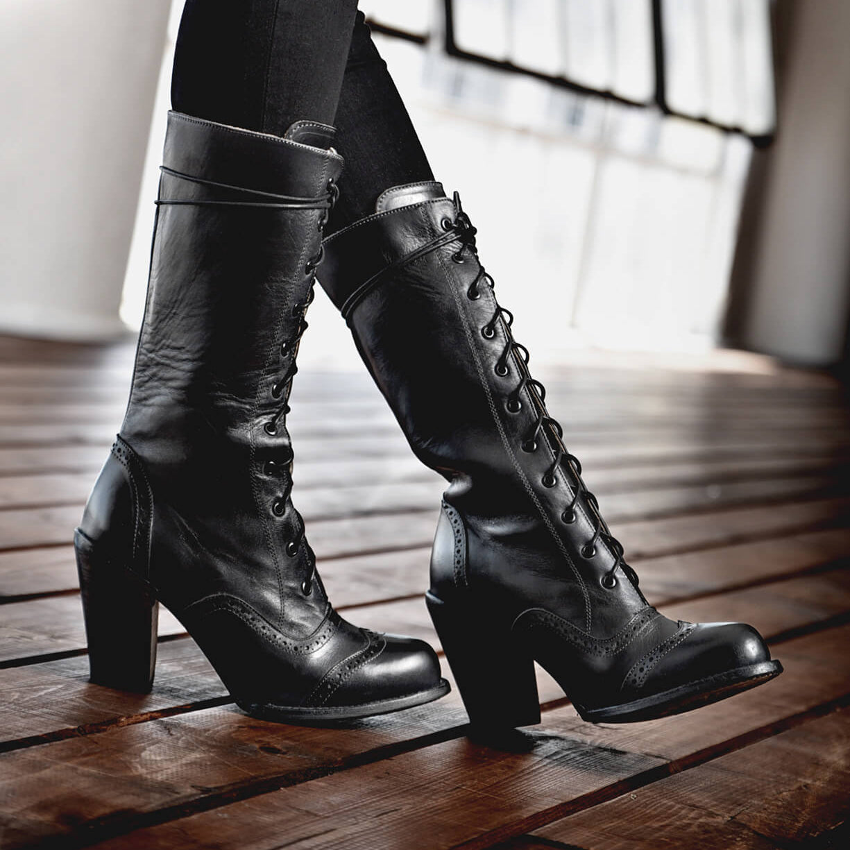 A woman wearing the Ariana black leather boots with hand-tooled pinking detail on a wooden floor.
