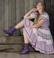 A woman in a Victorian style purple dress sitting on a wooden bench, showcasing uncompromising quality and hand dyed elegance, wearing the Eleanor dress by Oak Tree Farms.