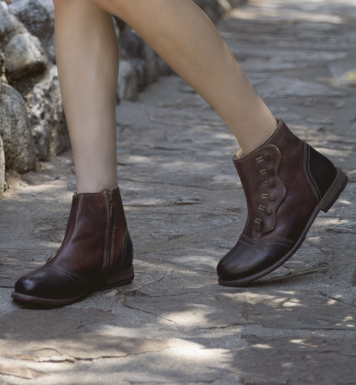 A woman wearing brown leather Josephine boots from Oak Tree Farms with a YKK zipper walking on a stone path.