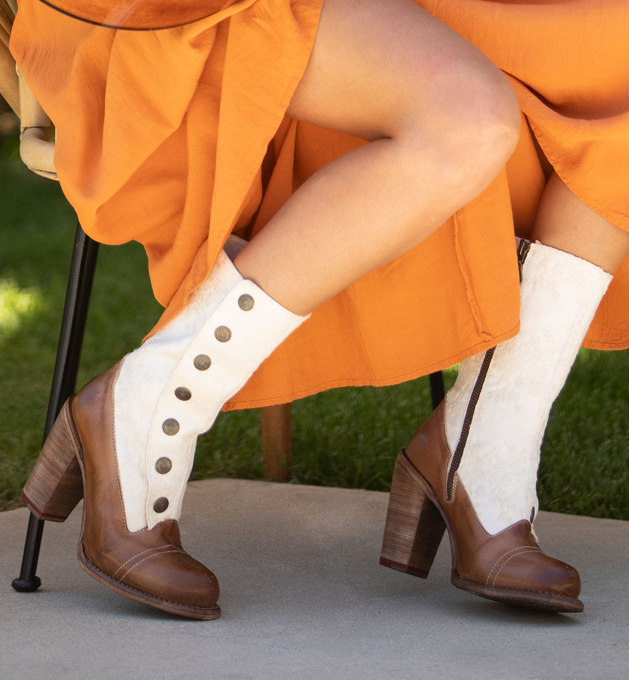 A woman in an orange dress is sitting on a bench wearing white and tan Amelia boots made by Oak Tree Farms, which are made of full grain leather.