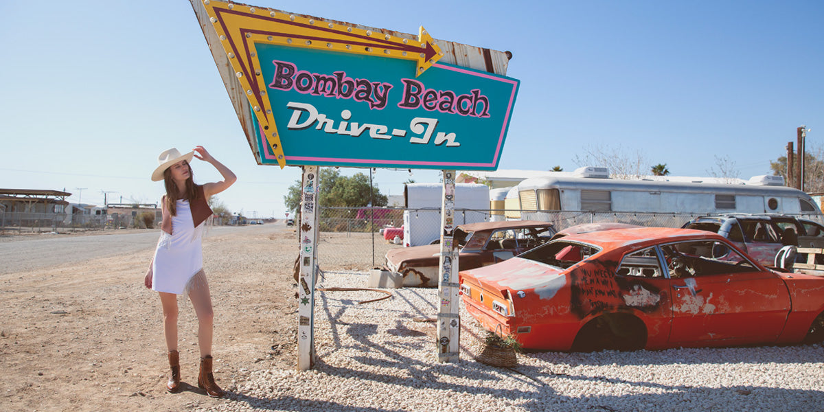 A woman in a white dress standing next to a car in a desert.