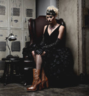 A woman in a black dress, wearing Oak Tree Farms' Amelia Steampunk boots made of full grain leather, sitting on a chair.