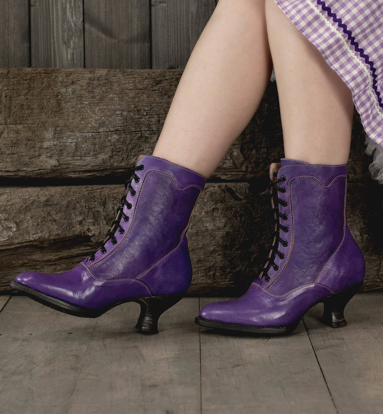 A woman displaying uncompromising quality in her Eleanor Victorian-style, hand-dyed purple leather boots as she stands on a wooden floor by Oak Tree Farms.