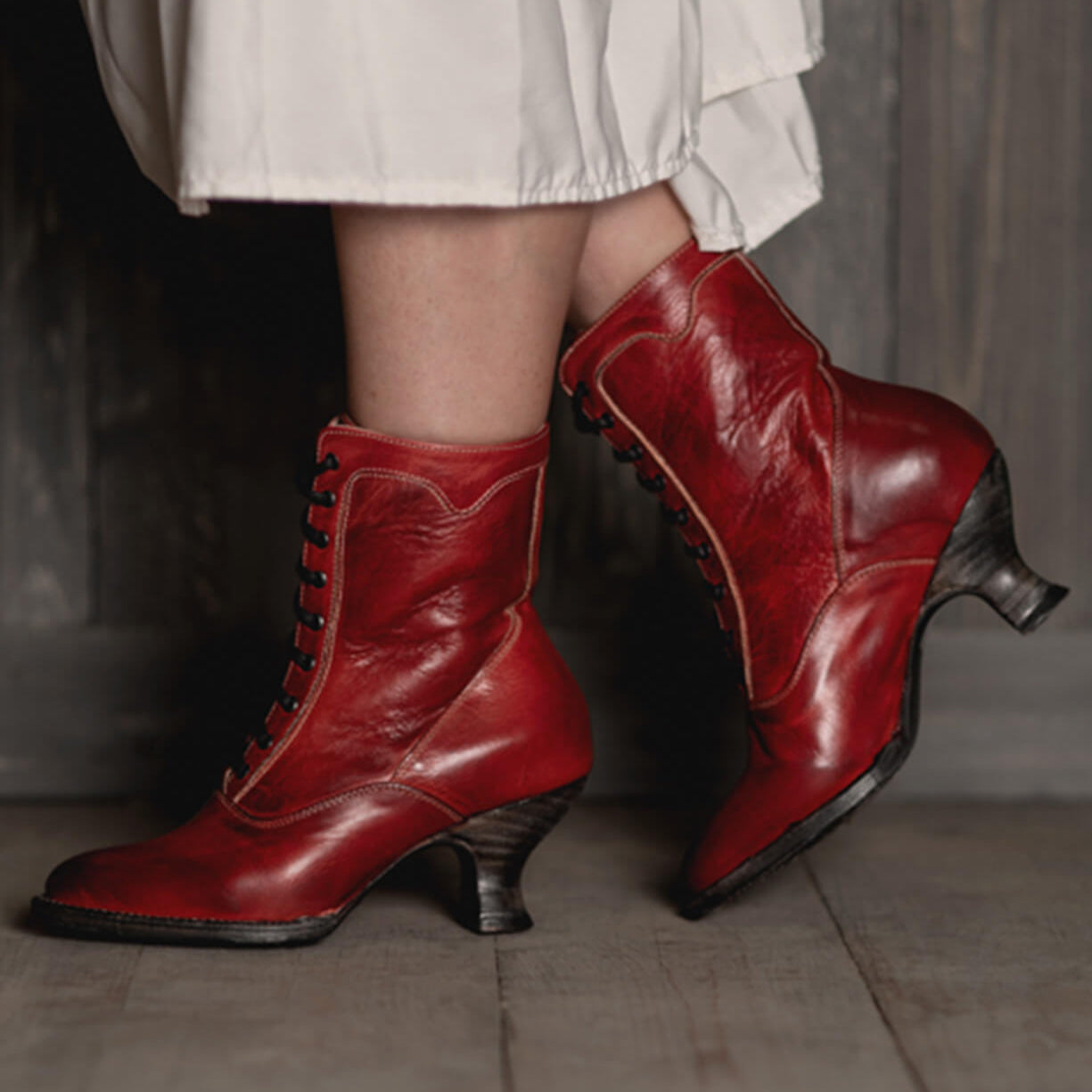 A woman with uncompromising quality, wearing Oak Tree Farms' Eleanor Victorian style hand dyed red leather boots, gracefully walks on a wooden floor.