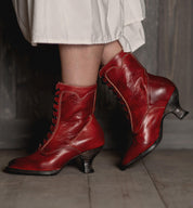 A woman with uncompromising quality, wearing Oak Tree Farms' Eleanor Victorian style hand dyed red leather boots, gracefully walks on a wooden floor.