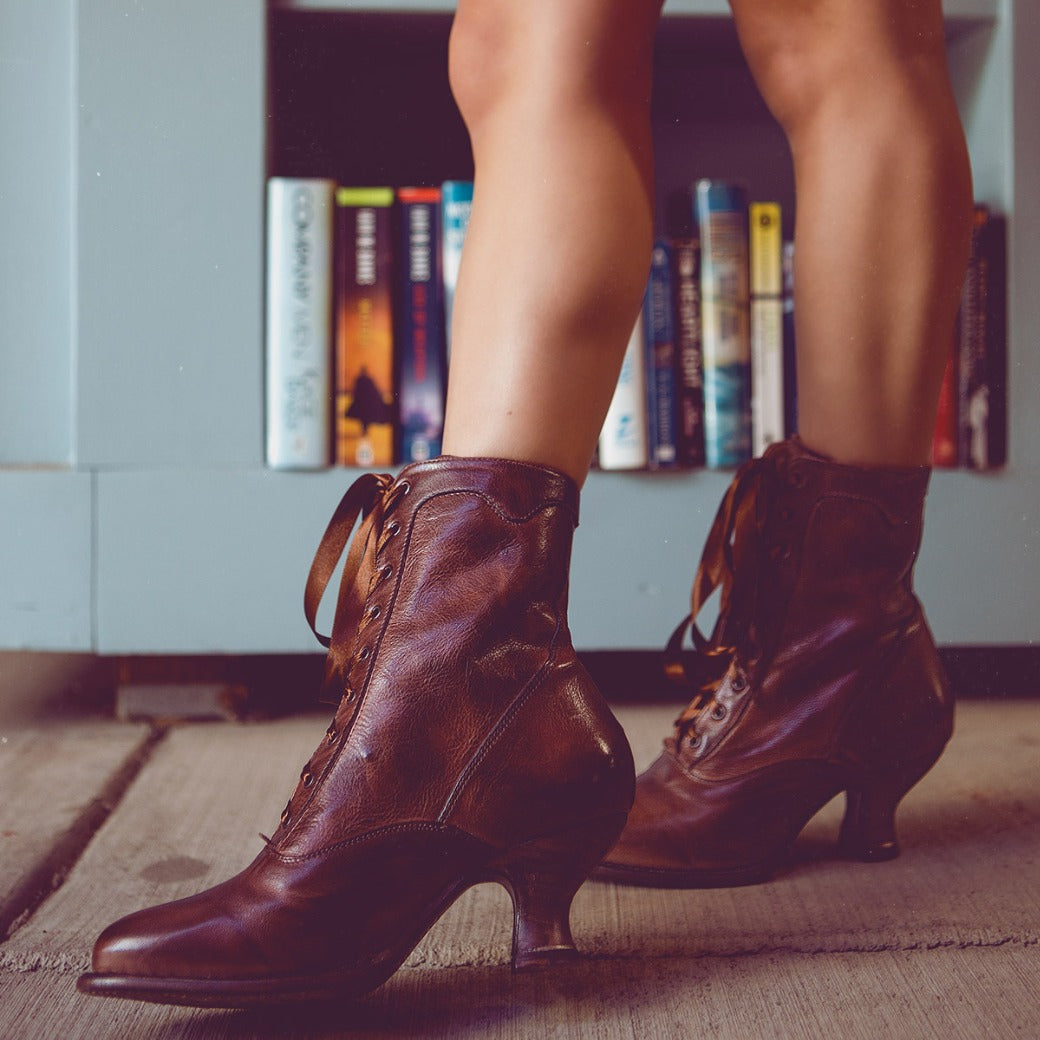 A woman wearing Oak Tree Farms' Eleanor hand-dyed brown leather boots in front of a bookcase, showcasing uncompromising quality.