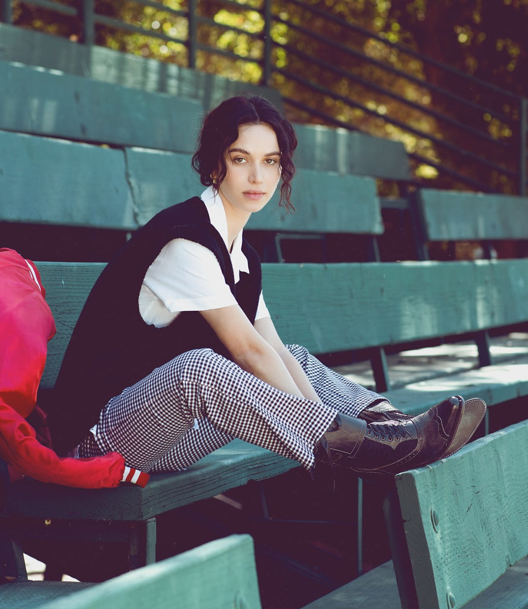 A woman sitting on green bleachers with a red Faye bag and wearing Oak Tree Farms handcrafted leather boots.