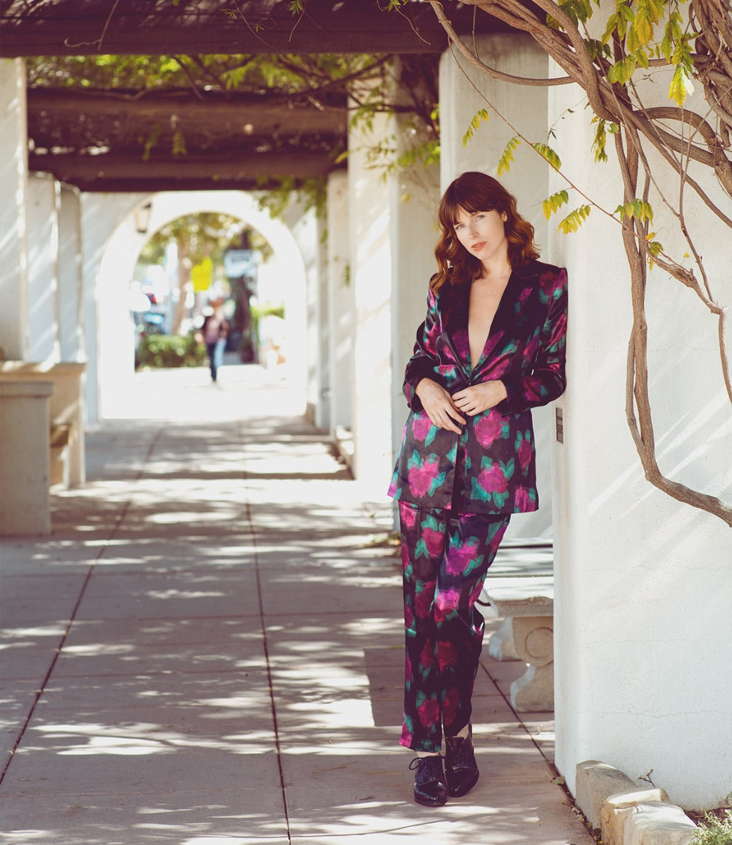A woman in a neutral look and floral suit, leaning against a wall, wearing the Janet suit by Oak Tree Farms.