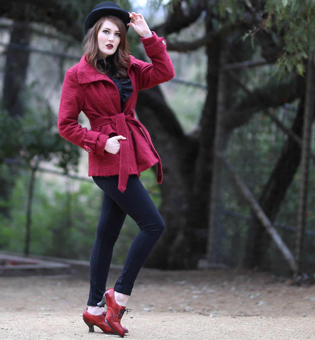 A woman in a red coat with a neutral look, posing in a wooded area wearing Oak Tree Farms leather shoes with lace-up front, named Janet.