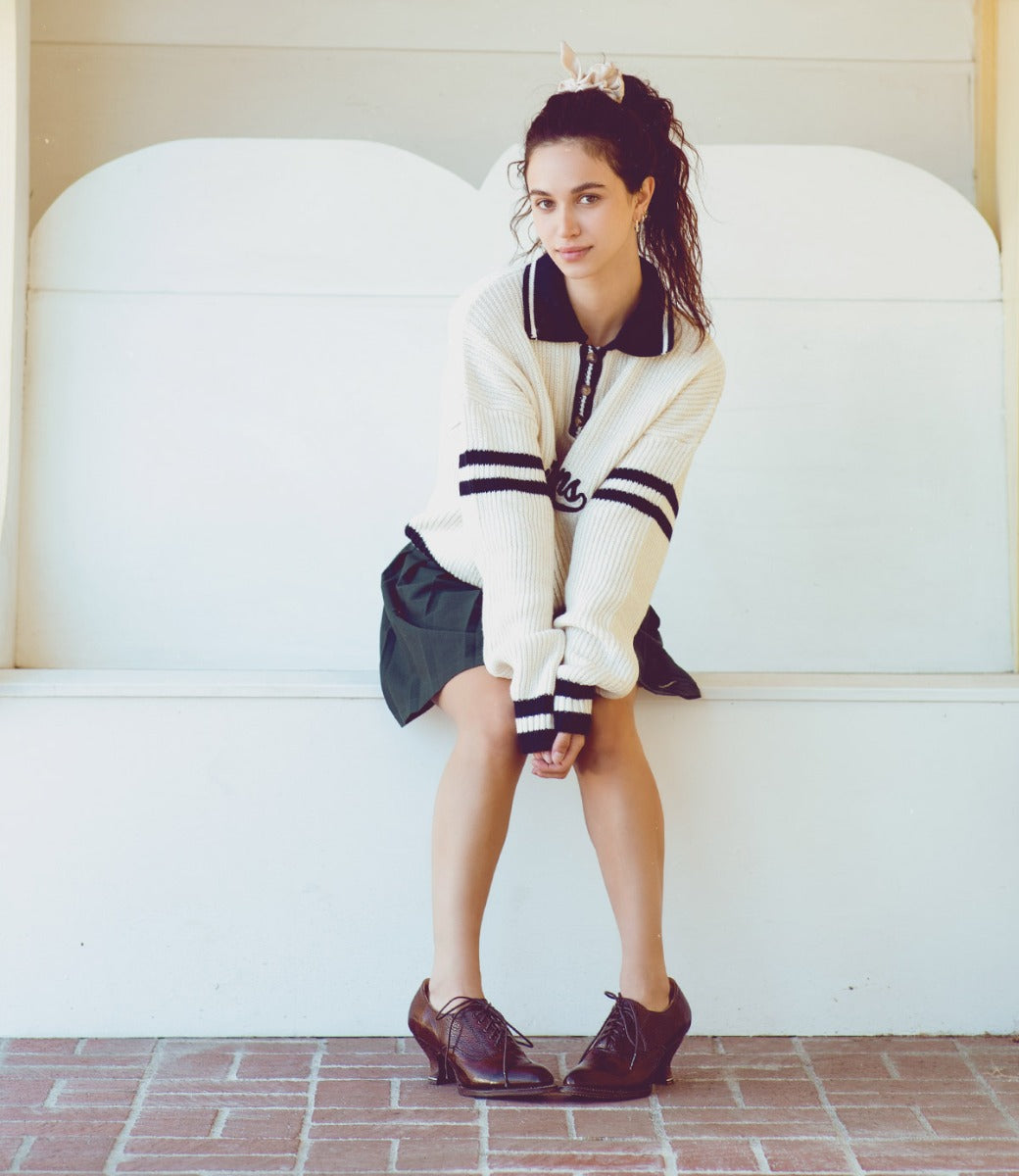 A girl sitting on a bench wearing a sweater and skirt, completing her neutral look with Oak Tree Farms lace-up front leather shoes named Janet.