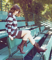 A woman, dressed in leather boots and hand dyed Oak Tree Farms Black Rustic clothing, is sitting gracefully on a green bench.