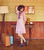 A woman in a pink dress standing next to a Mini suitcase, exuding both comfort and stacked heeled beauty from Oak Tree Farms.